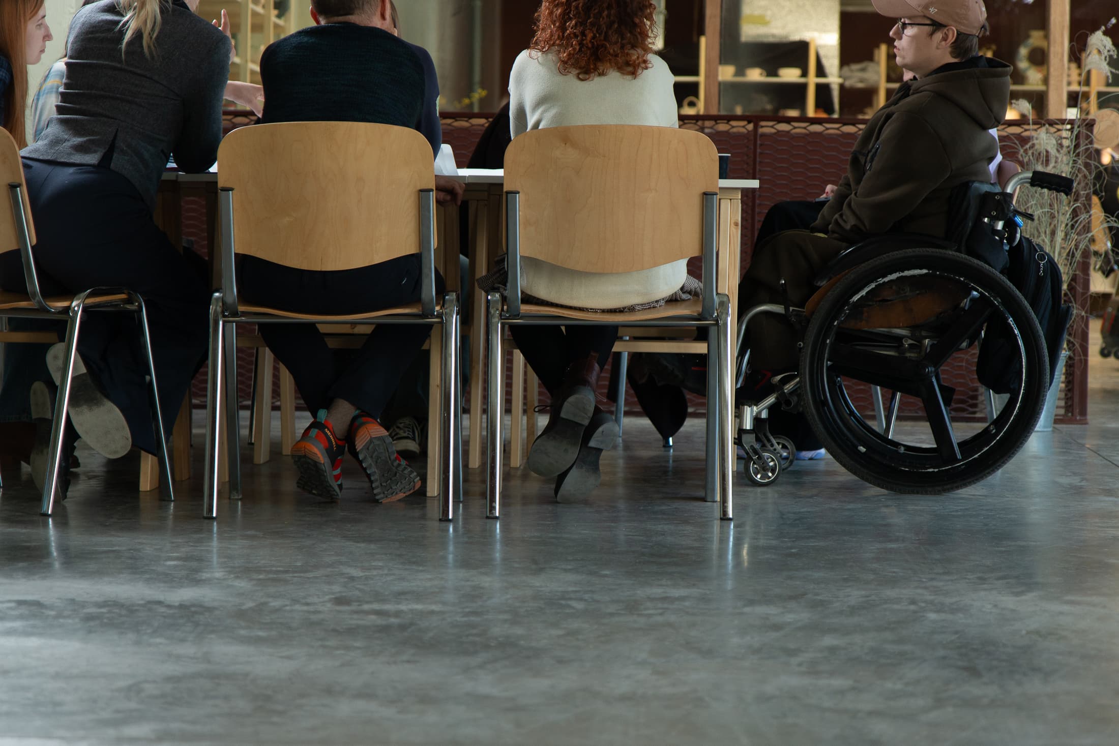 A snapshot of a working moment: four people are sitting on wooden chairs with their backs to the camera, while the fifth participant, wearing a brown hoodie and cap, is sitting in a wheelchair at the right edge of the table. They are focused on their shared task. The shot is taken from a low angle, emphasising the equality of all participants at the table and the inclusiveness of the workspace.