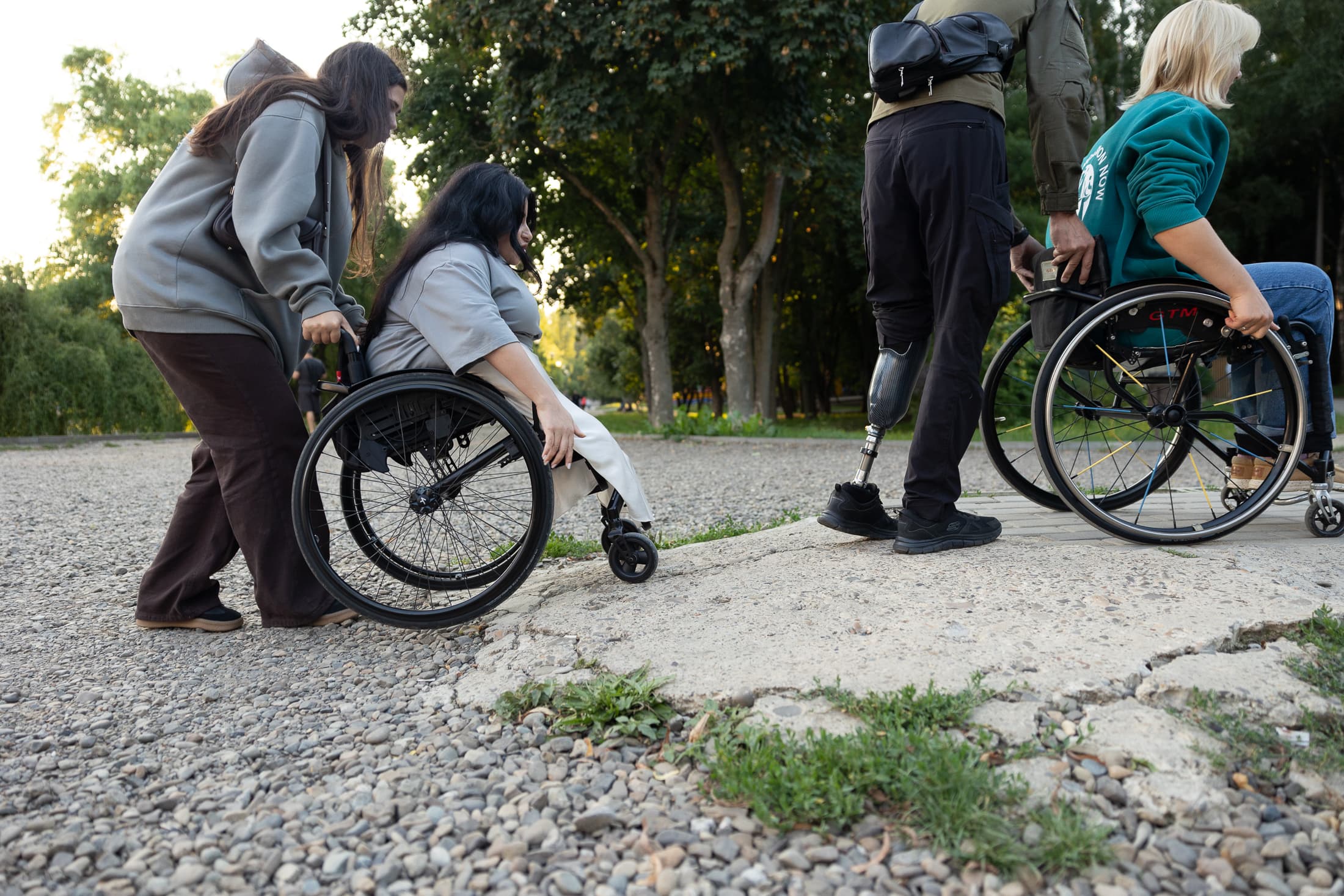 A photograph of a walk in the park illustrating a lack of accessibility. In the foreground, a woman is helping another woman in a wheelchair to navigate a section of deep gravel and broken concrete. A man with a prosthetic lower limb walks nearby, and another person in a wheelchair walks ahead. The contrast between the green trees and the damaged road highlights the problem of physical barriers in recreational areas.