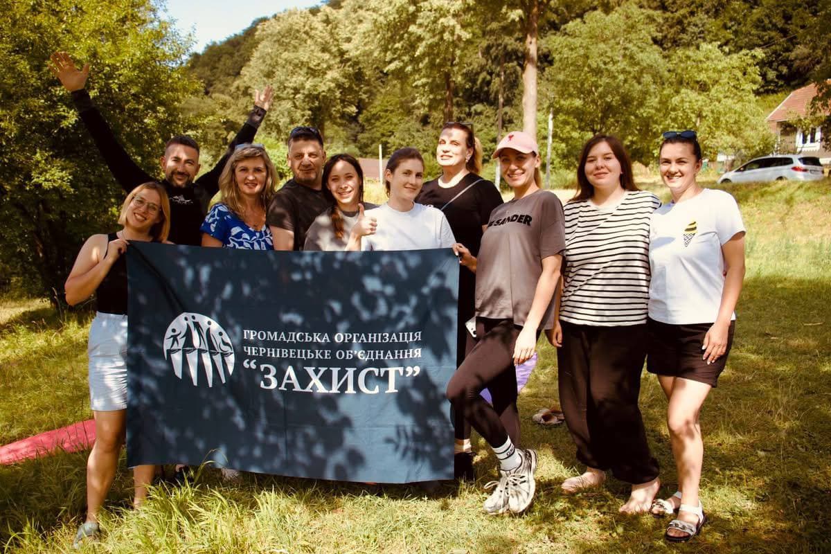 A sunny group photo of ten young people in a forest glade. The team stands in two rows, smiling and hugging each other. In front, two girls hold an unfurled dark blue flag of the ZAHYST organisation with a logo featuring silhouettes of people holding hands. In the background are dense green trees and a hillside. The atmosphere is friendly, summery and full of energy.