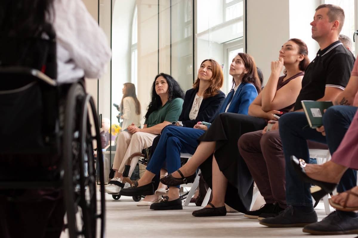 Крупний план аудиторії у світлому сучасному залі. Кілька жінок та чоловік сидять у ряду, дивлячись угору на спікера або екран із виразом зацікавленості та роздумів. Зліва в кадрі видно частину крісла колісного та спину учасника, який перебуває в центрі подій.