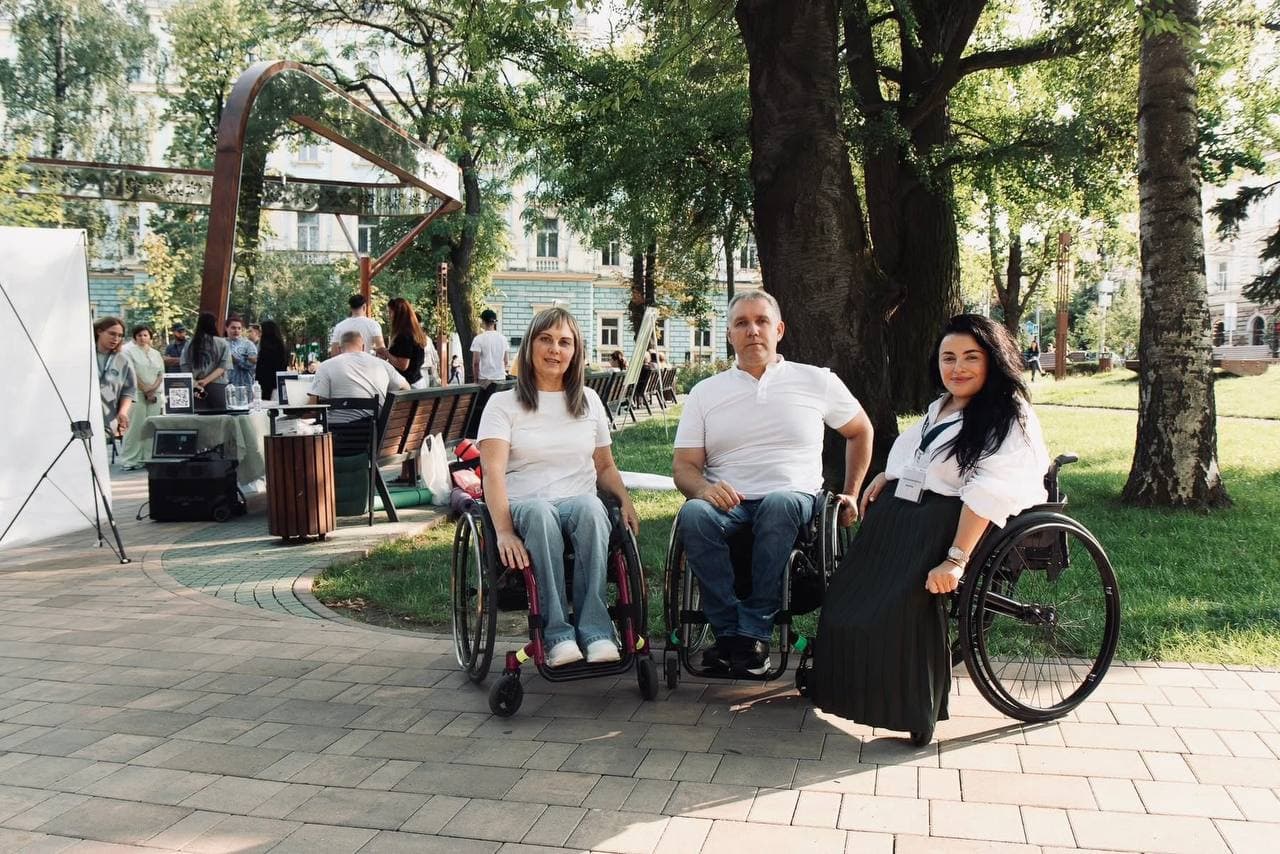 Групове фото трьох активістів на фоні міського парку у світлу пору доби. У центрі — чоловік у білій футболці та джинсах, ліворуч — жінка у білій футболці та світлих штанах, праворуч — жінка у білій блузі та довгій чорній спідниці. Усі троє сидять у кріслах колісних на рівній брукованій поверхні, що демонструє доступність простору. На задньому плані ліворуч розташована сучасна дзеркальна архітектурна конструкція, а праворуч — газон та дерева.