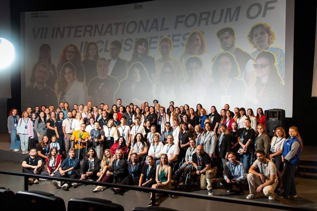 Масштабне спільне фото понад 90 учасників та організаторів форуму в темному залі з професійним освітленням. У центрі перших рядів сидять люди у кріслах колісних, поруч із ними — волонтери та експерти. Багато учасників мають сині стрічки з бейджами на шиї. Позаду групи височіє екран із написом «VII INTERNATIONAL FORUM OF INCLUSIVENESS» та великими портретами спікерів у жовтих рамках. Кадр передає атмосферу великої та впливової спільноти.