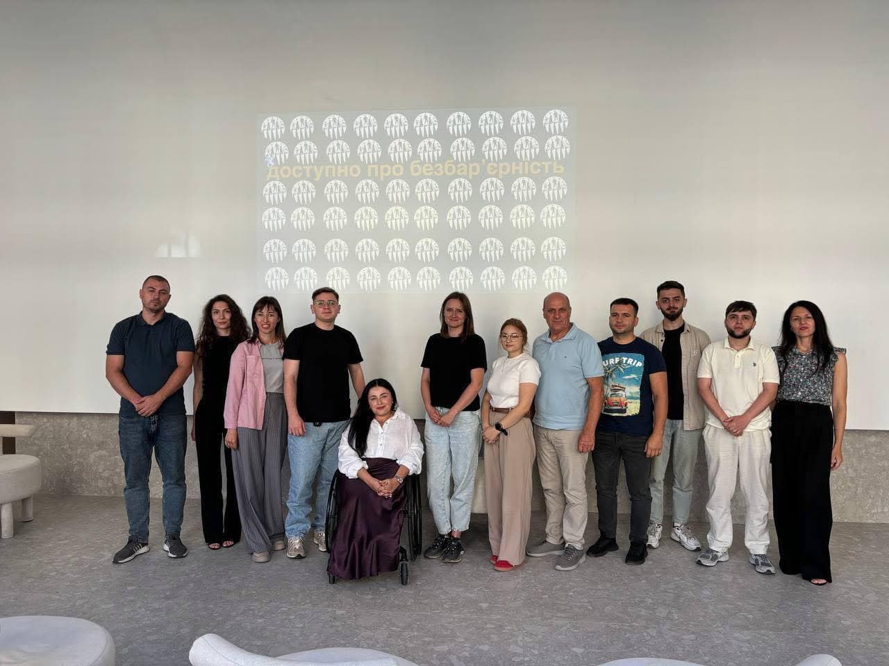 Спільне фото одинадцяти людей у світлому залі. Учасники стоять у ряд, а в центрі композиції сидить жінка у білій блузі та довгій темній спідниці у кріслі колісному. На стіні позаду них за допомогою проєктора виведено напис «Доступно про безбар'єрність» та патерн із білих круглих логотипів організації.