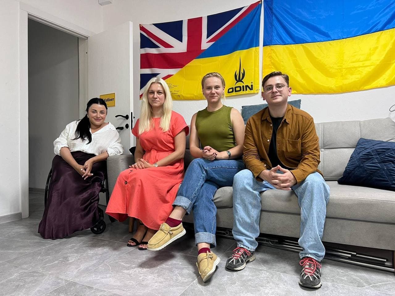 Четверо учасників зустрічі (три жінки та чоловік) сидять на сірому дивані в офісному приміщенні. На стіні позаду них висять прапори України та Великої Британії.
