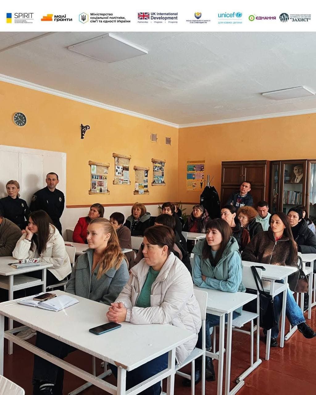 A photograph of a social project in action in the Chernivtsi region. About 20 people have gathered in a bright classroom with orange walls. In the foreground, women are sitting at white desks, some of them taking notes. On the left, near the door, two uniformed police officers are standing, also attending the meeting. At the top of the photo is a row of partner logos, including SPIRIT, UNICEF, and the Ministry of Social Policy of Ukraine.