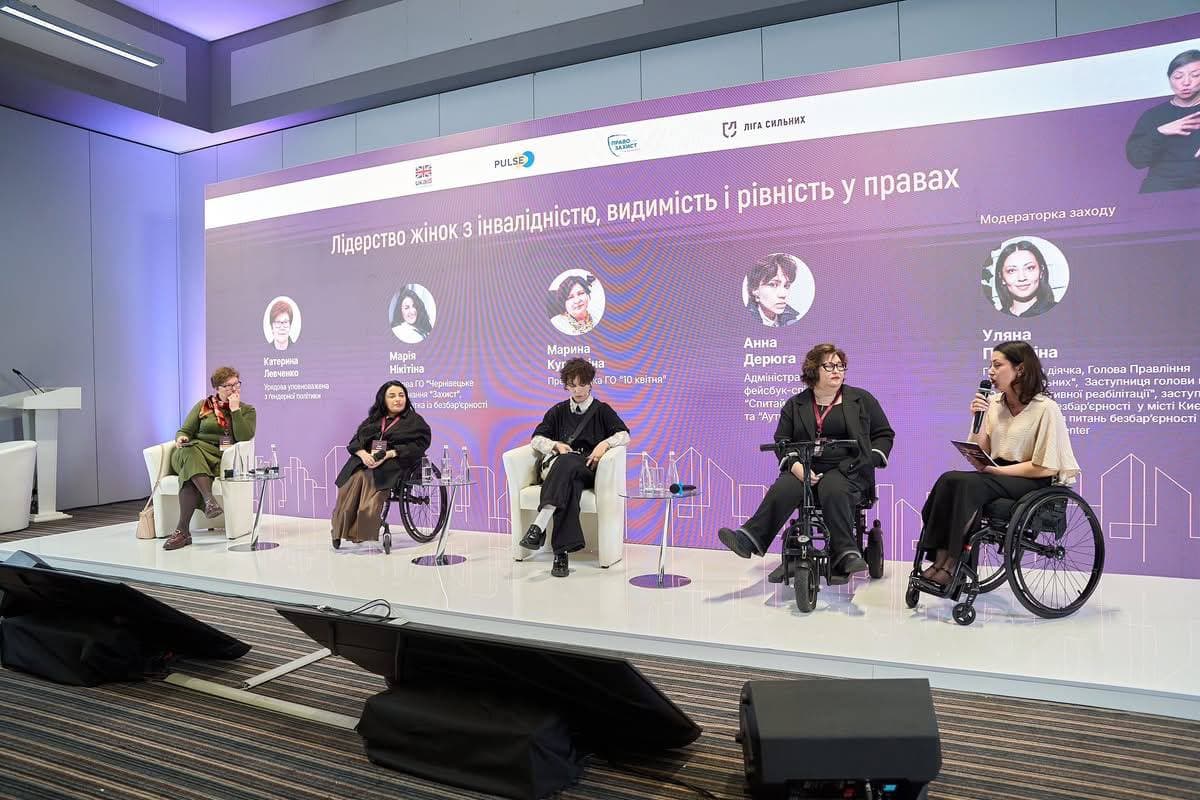 Сцена Національного форуму з великим мультимедійним екраном, на якому відображені імена та фото спікерок дискусійної панелі. На екрані напис: «Лідерство жінок з інвалідністю, видимість і рівність у правах». У кріслах на сцені сидять учасниці, зокрема Катерина Левченко, Марія Нікітіна, Марина Кучміна та Анна Дерюга. Праворуч модераторка заходу Уляна Пчолкіна, сидячи у кріслі колісному, звертається до аудиторії з мікрофоном. У верхньому правому куті екрана видно вікно з перекладачкою жестової мови.