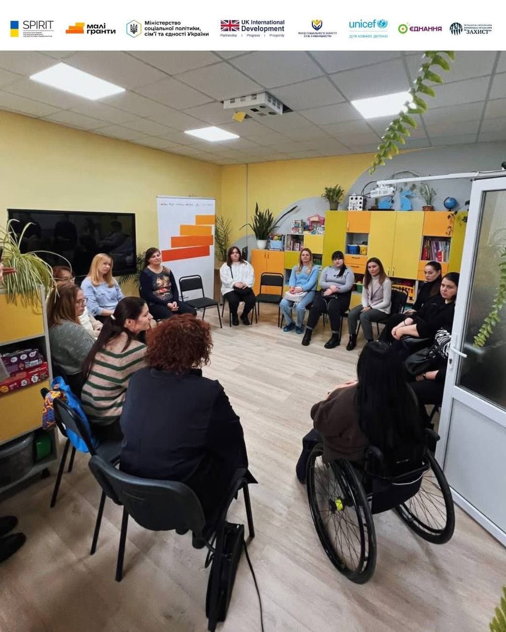 A group of about 12 women gathered for a discussion in a bright room resembling a children's center or kindergarten group. The participants are seated on chairs arranged in a circle. In the foreground, a woman in a wheelchair, with her back to the camera, listens to the other participants. Yellow toy cabinets, a globe, and indoor plants are visible in the interior.
