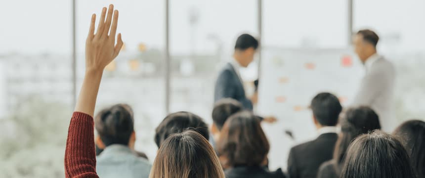 In the foreground on the left, you can clearly see the raised right hand of a person in a burgundy sweater. The rest of the frame is out of focus: you can see the backs of the heads of other listeners and two speakers in business suits standing next to a flipchart with colored stickers. The bright room with panoramic windows creates a professional and open atmosphere of a business workshop.
