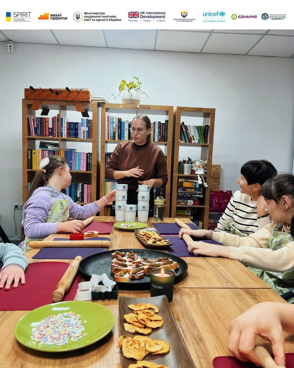 The participants of the social event gathered around a table in a room with large bookshelves. The hostess, wearing glasses and a brown sweater, stands in the center, stacking white paper cups into a tower. In front of the girls on the table are rolling pins, work stands, plates with snacks (cucumber sandwiches, chips) and sprinkles for desserts. The atmosphere is focused and cozy, with a decorative candle burning on the table.
