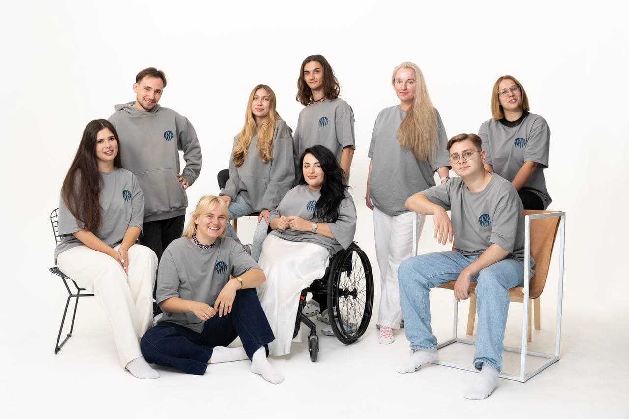Колективне фото на білому фоні: п'ятеро людей стоять на задньому плані, троє сидять на стільцях та підлозі, а одна жінка розташована в центрі на кріслі колісному. Всі одягнені в корпоративний сірий мерч. Світле освітлення та відкриті пози створюють позитивний образ команди.